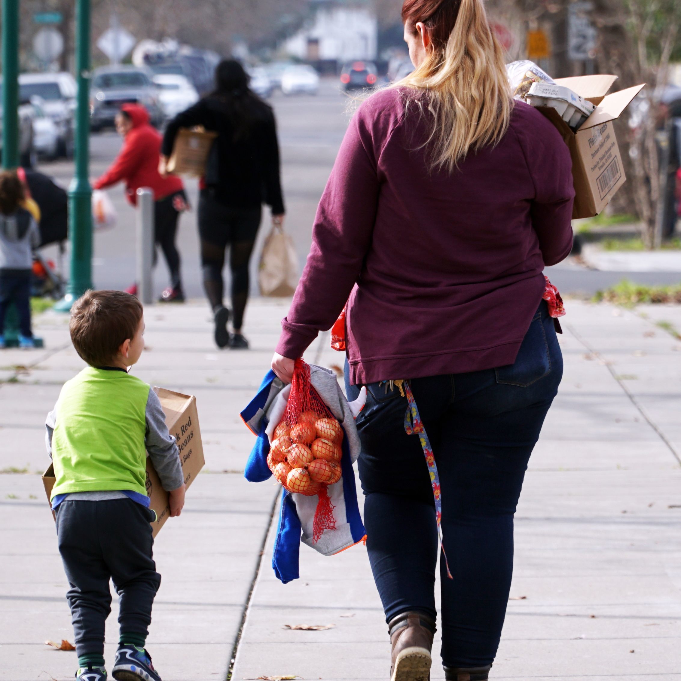 Back view of mother and son walking away from a distribution site carrying lots of groceries