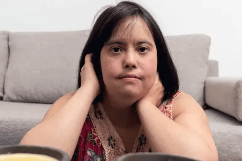 A woman with short dark hair is sitting on the floor in front of a beige sofa. She is leaning on a surface with her hands near her face. She is wearing a colorful patterned sleeveless top. Two out-of-focus cups or bowls are visible in the foreground.