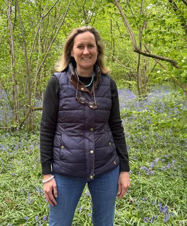 Tall woman with light brown hair, vest and blue jeans stands smiling at camera. She is in a wooded area with a stream running behind her. 