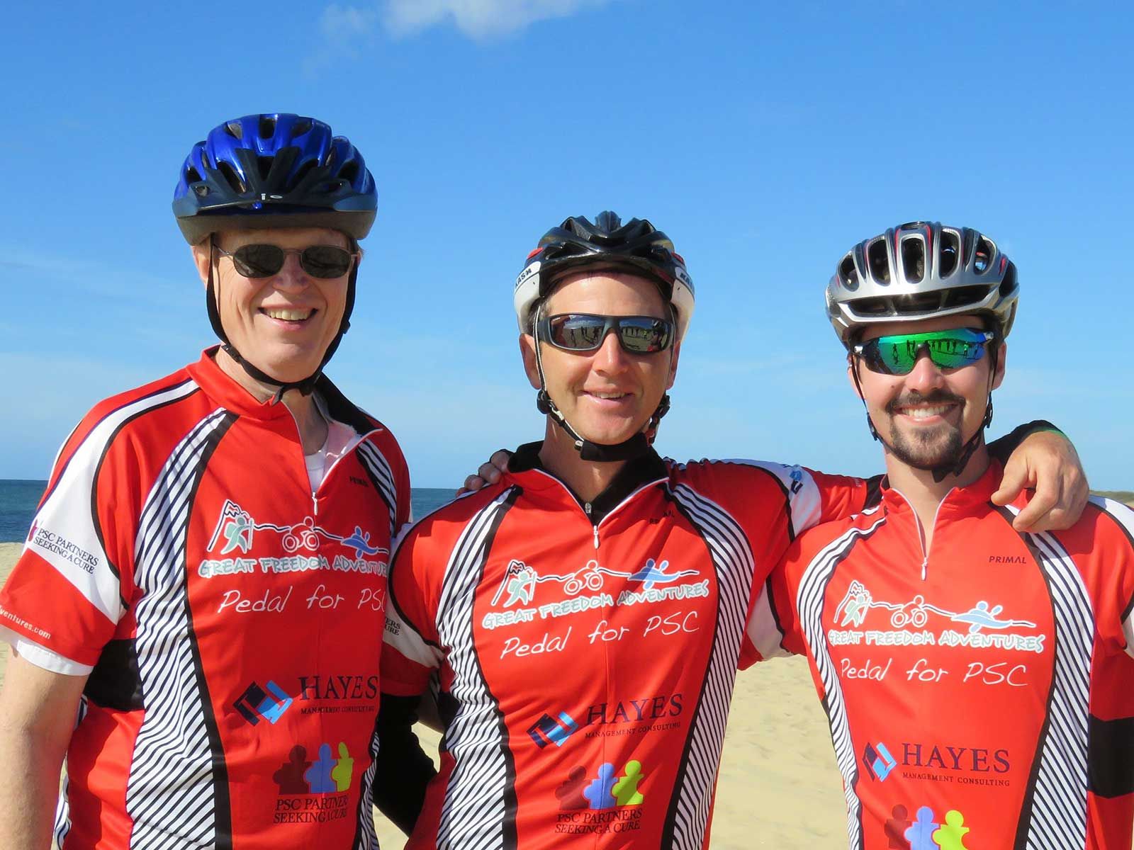 Three men in bicycle helmets stand in a line with their arms around each other. Their shirts read Pedal for PSC