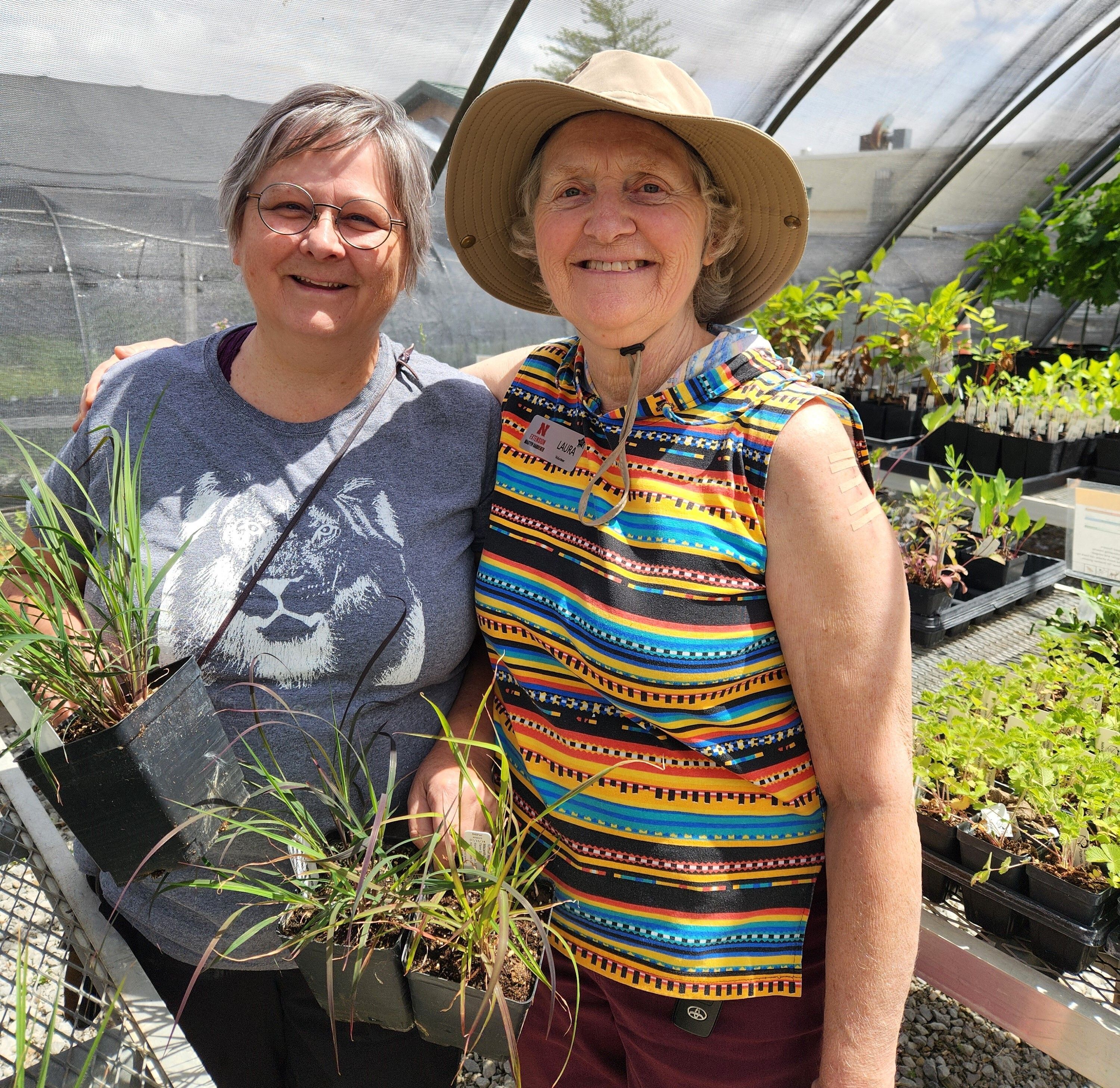 A woman wearing a gray t-shirt stands with a woman wearing a multi-color shirt and wide-brimmed hat in a greenhouse with plants. 