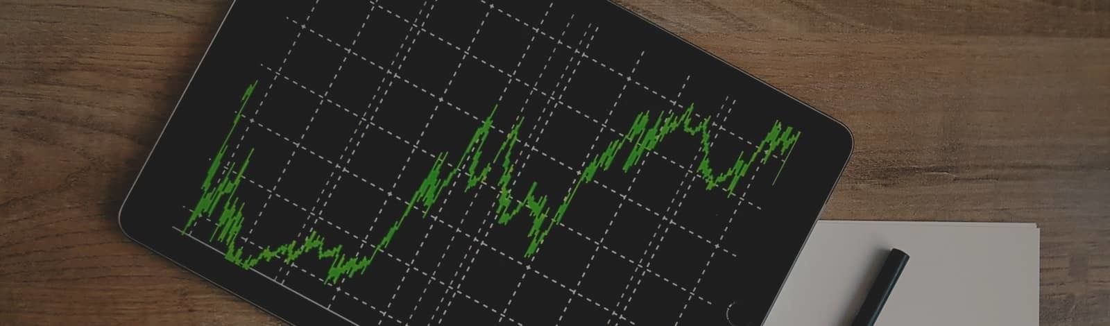 Areal view photo of a tablet set at a diagonal on a wooden table. The tablet has a black background with a grid of white dotted lines and a green line graph showing periodic growth as it moves from left to right. There is white blank paper and a black pen