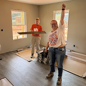 Volunteer raising a fist in celebration while another carries flooring material in a room with new laminate flooring