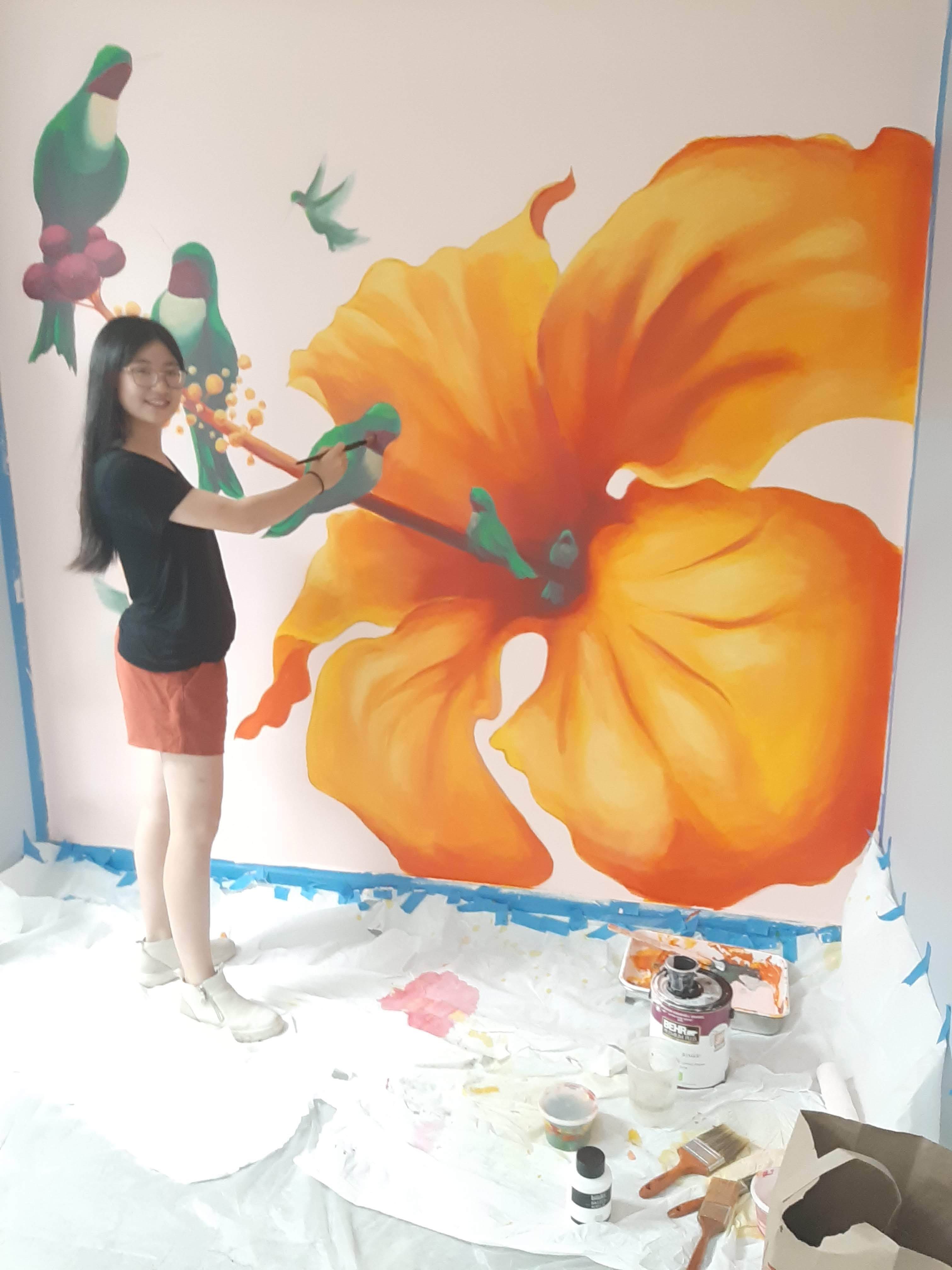 Shenendehowa student, Casey Gao painting orange hibiscus and hummingbird mural at Coburg Village fitness center