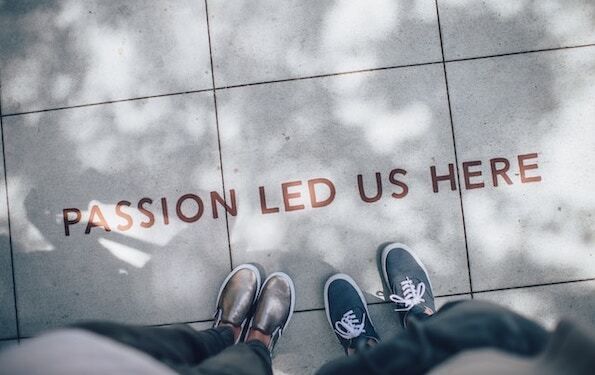 Aerial view photo looking down on a tiled sidewalk with the phrase "Passion Led Us Here" in brown letters from left to right. Two sets of sneakers peak out from the bottom, one is gold without laces and the other is navy blue with white laces. There is da