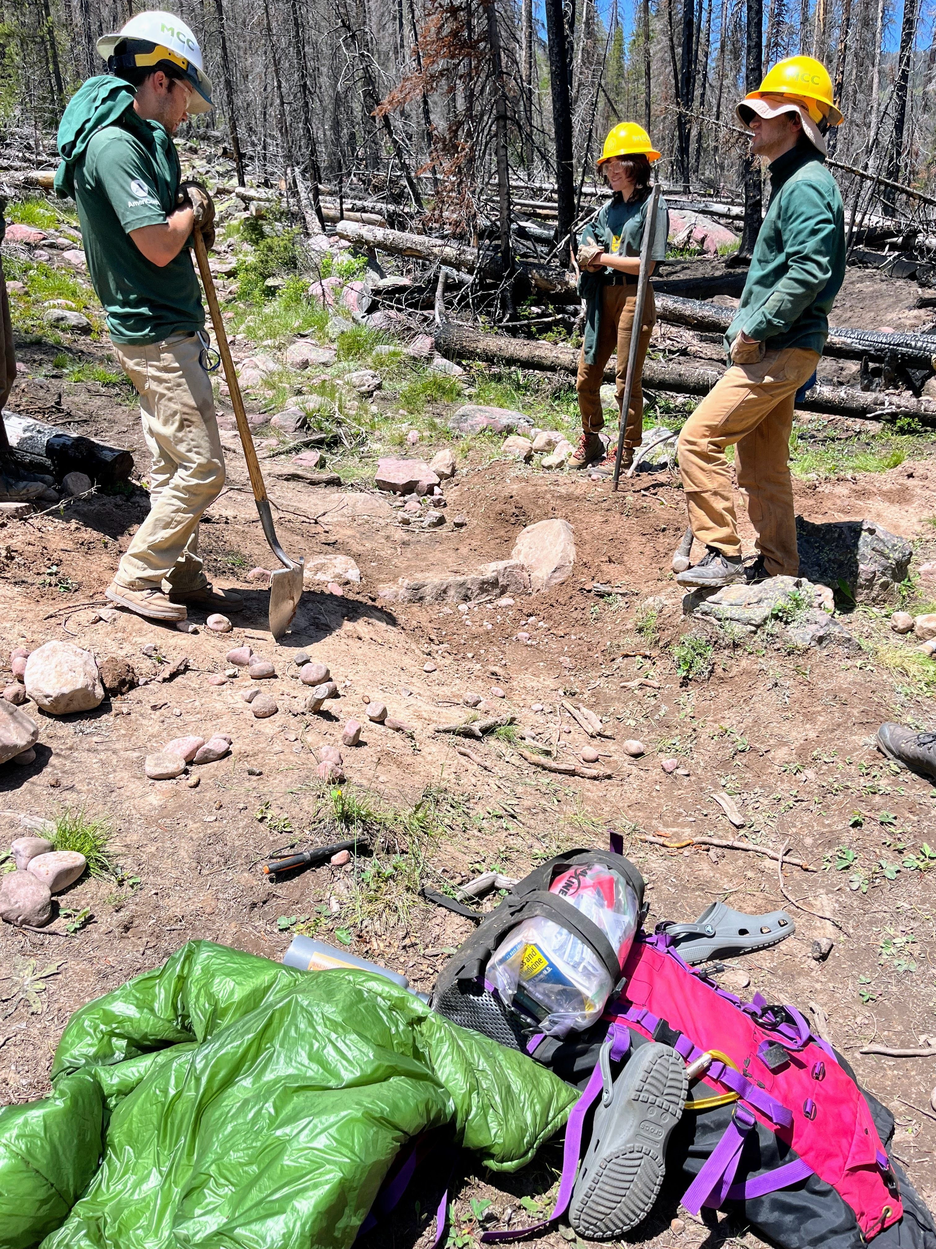 A crew stands in a circle holding rock bars and looking at rocks buried under the trail to create water drainage.