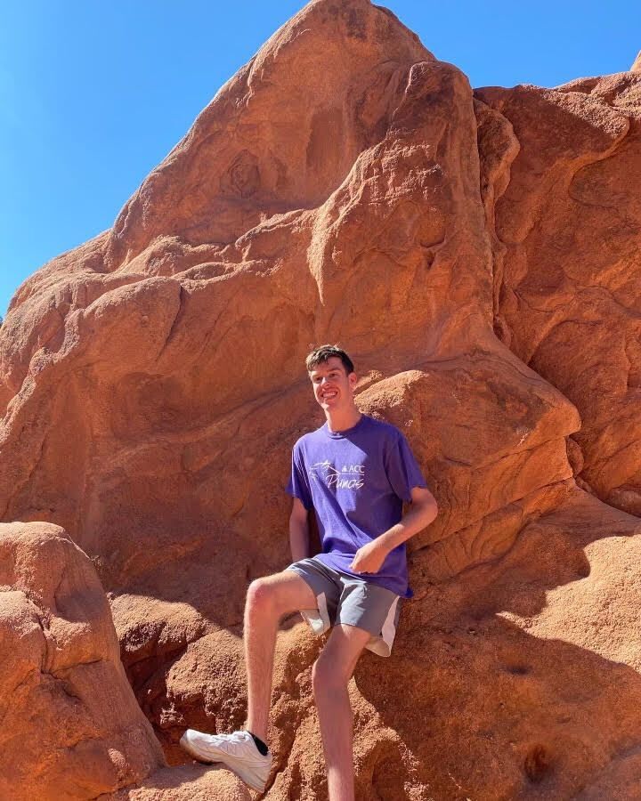 Bo wearing a purple ACC shirt while sitting on a big rock and smiling 