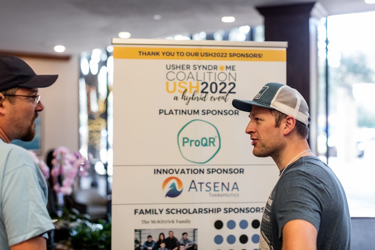 A young man wearing a hat is standing in front of a banner that has the Usher Syndrome Coalition logo as well as all of the sponsors this year.