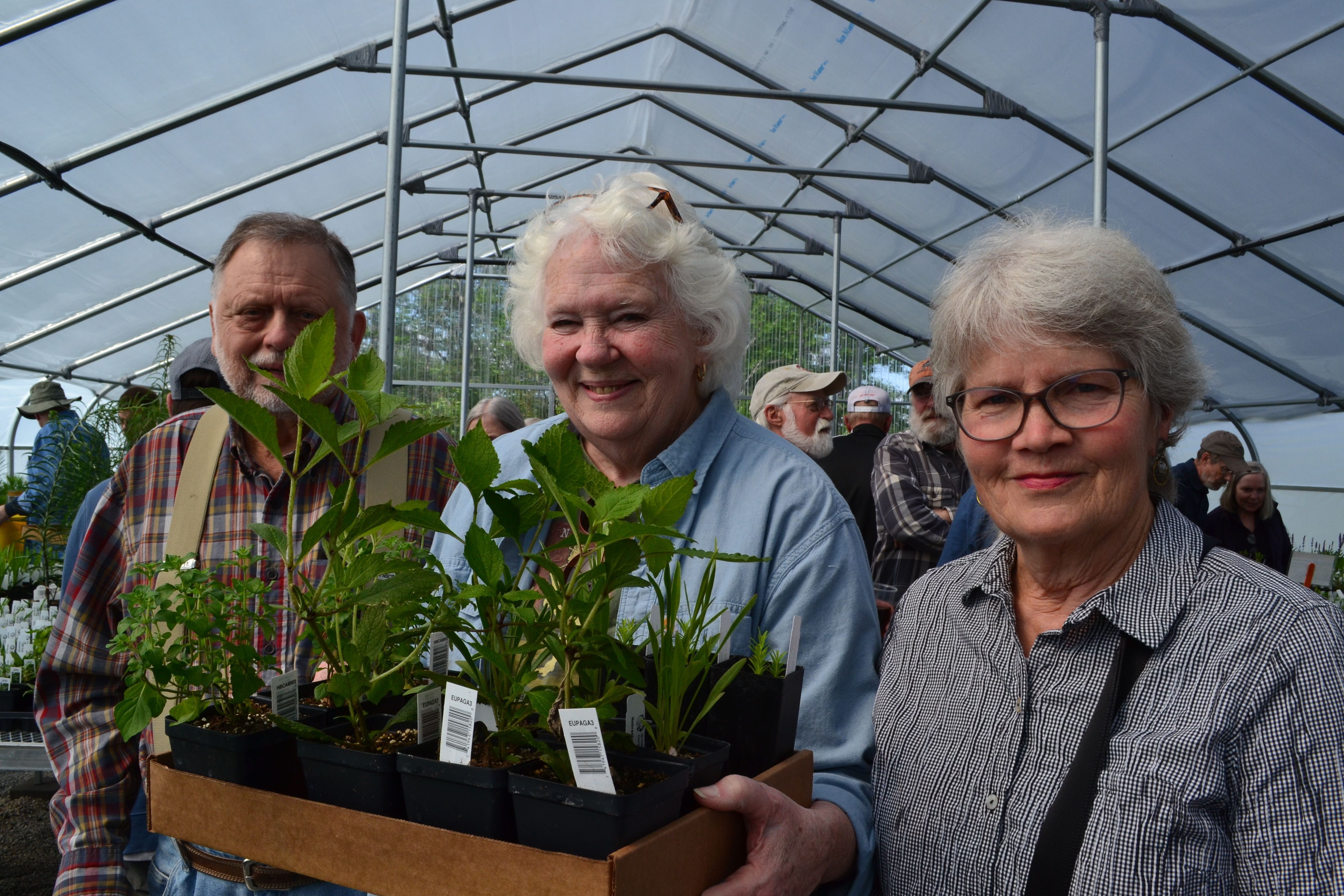 A man in a plaid shirt, a woman in light blue denim shirt and a woman in a gray shirt stand side-by-side in a greenhouse holding a box of plants. 