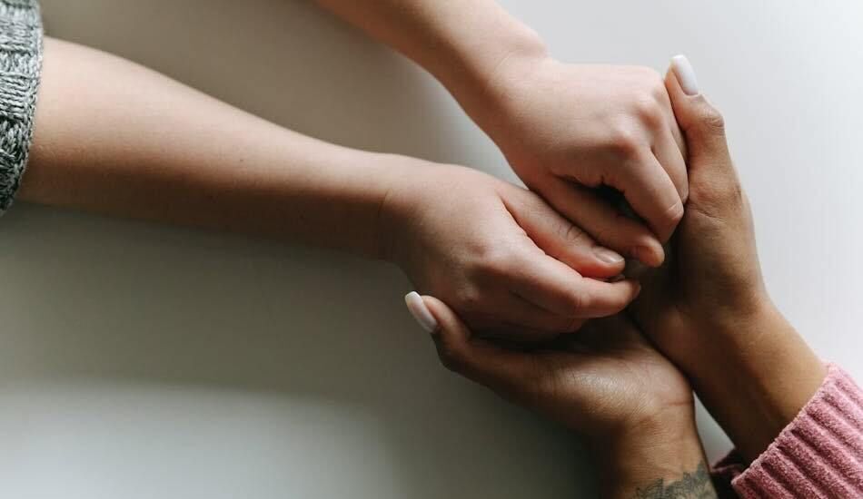 Photo of an adult holding lovingly the hands of a child, from underneath. Their hands sit atop an off-white tabletop.