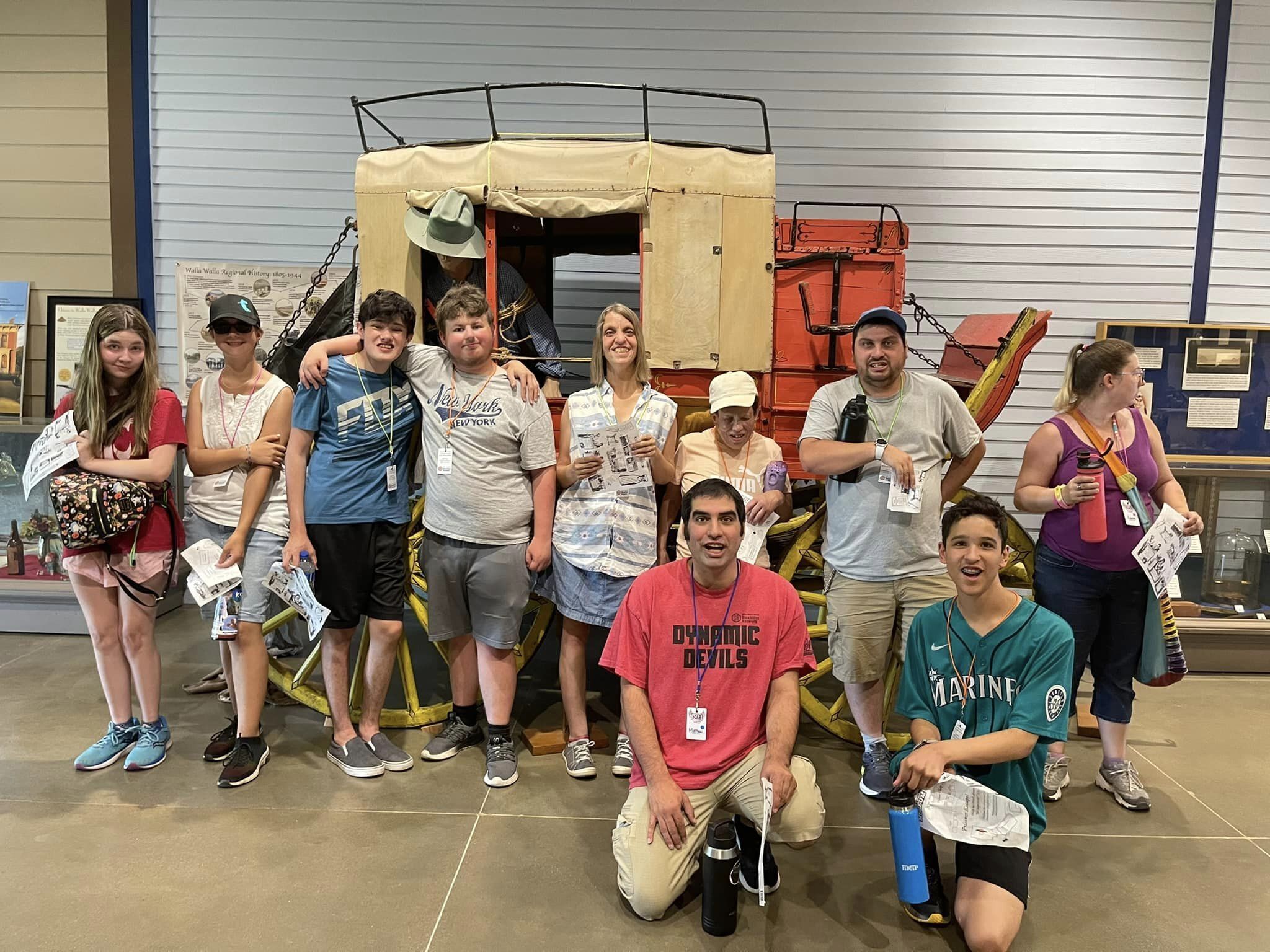 Group of teens and young adults with disabilities standing in front of a wooden stagecoach