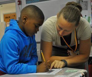 boy at a desk writing with a pencil and teacher with him