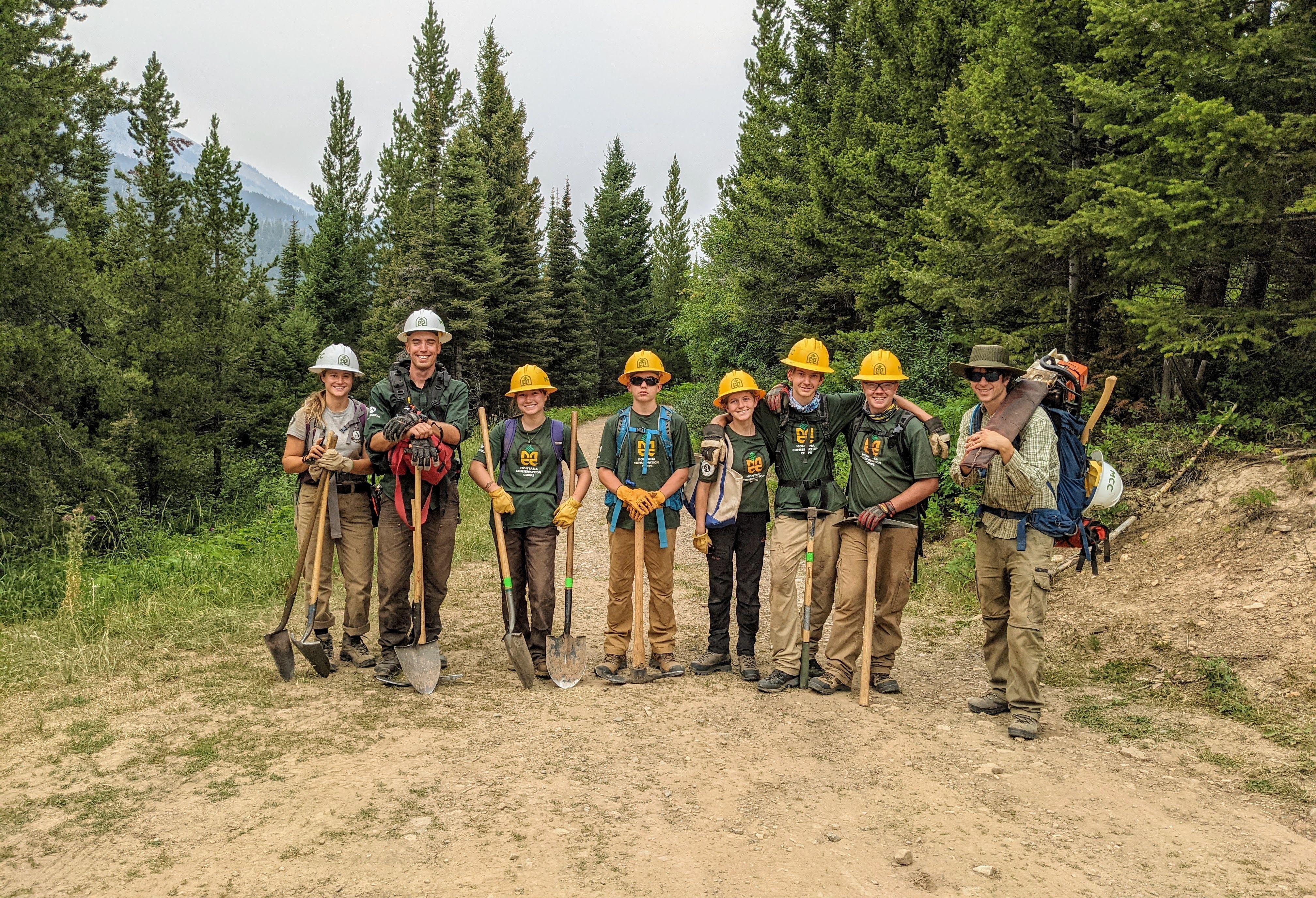 [Image Description: Eight MCC members standing together on a trail wearing their uniforms and helmets, holding shovels and pulaskis. Five of the members are youth, standing alongside their expedition leaders.]