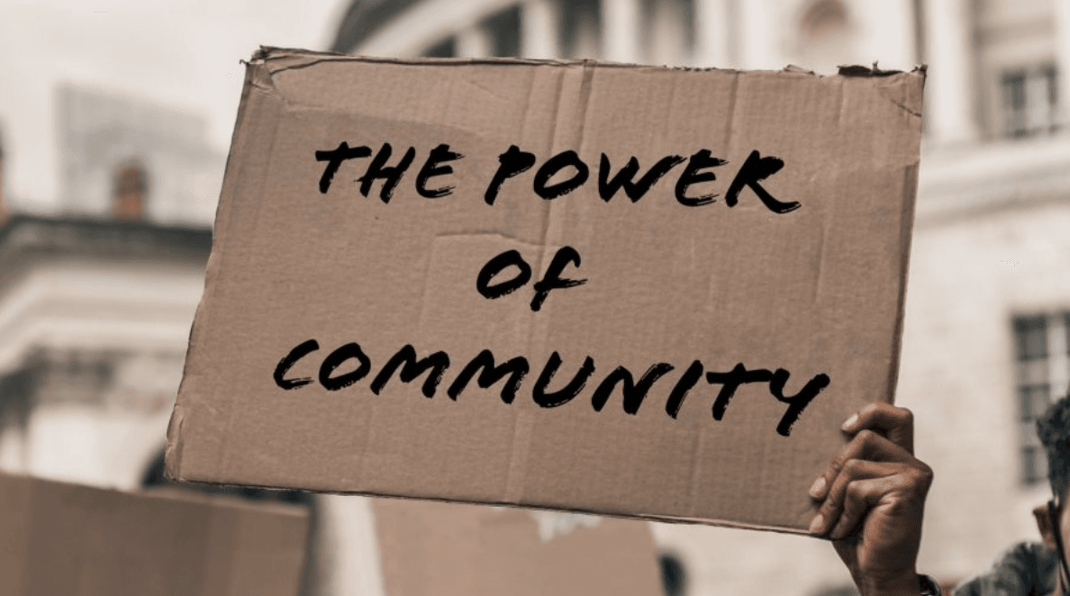 A hand holds a cardboard sign that reads in black ink: "THE POWER OF COMMUNITY."