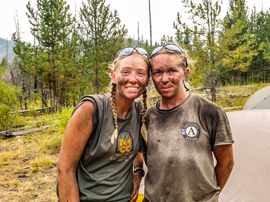 [Image Description: Two MCC Members standing side by side, smattered with dirt and smiling in a field with trees.]