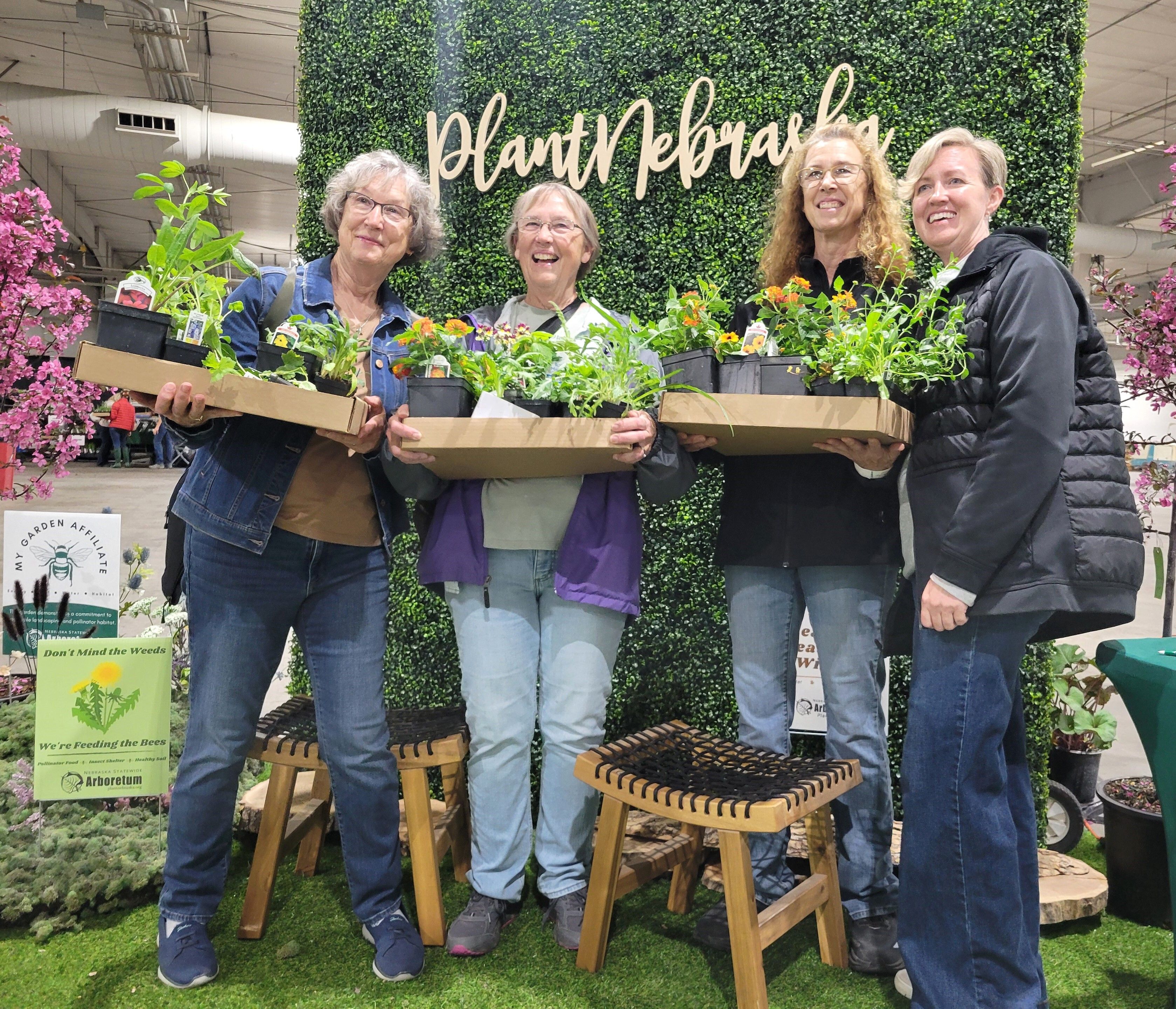 Four women holding flats of plants standing in front of the PlantNebraska sign on a green wall. 