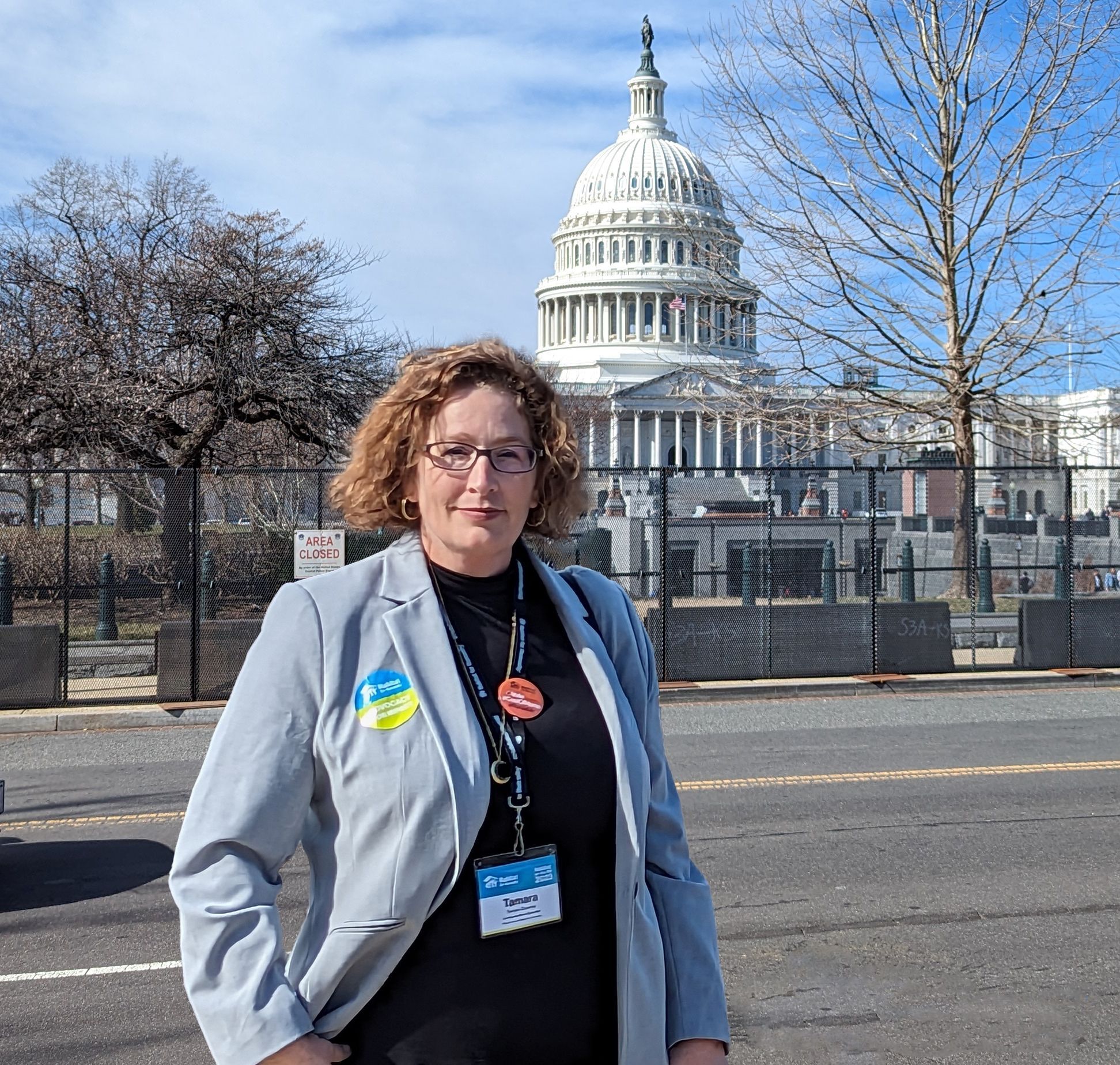 A person stands confidently before the United States Capitol Building. With a name badge and advocacy buttons, their poised demeanor suggests participation in a significant event related to legislative advocacy.