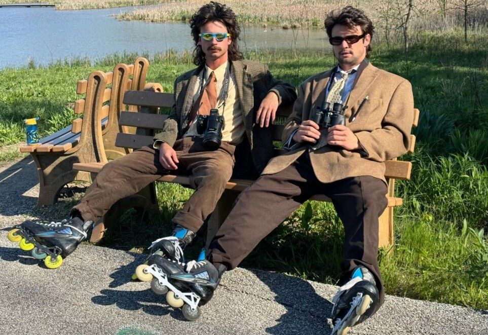 Two men sit on a park bench wearing roller blades, suits, and sunglasses. One has a DSLR camera and the other has binoculars. The vibe is very unserious. 