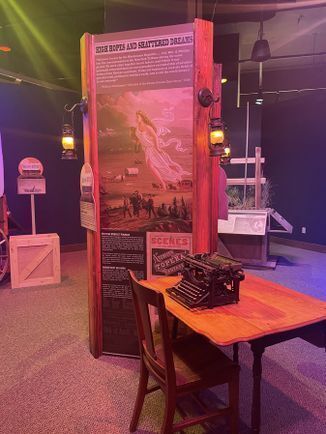 Exhibit panel, table with a typewriter, and chair for the Smoky Hill Museum exhibit "A Place to Call Home" on immigration into Kansas.