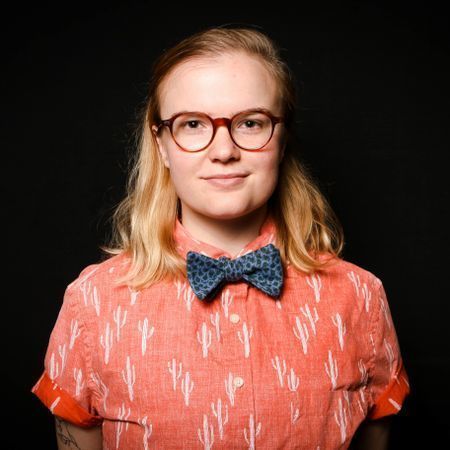 Morgan Allen-White's headshot. She has blonde hair and wears glasses. She wears a bright orange shirt and bowtie.