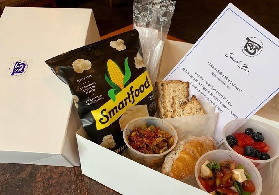 A photograph of a cardboard take out box containing a chicken salad croissant, tapenade, and other foods with a Trio's restaurant card