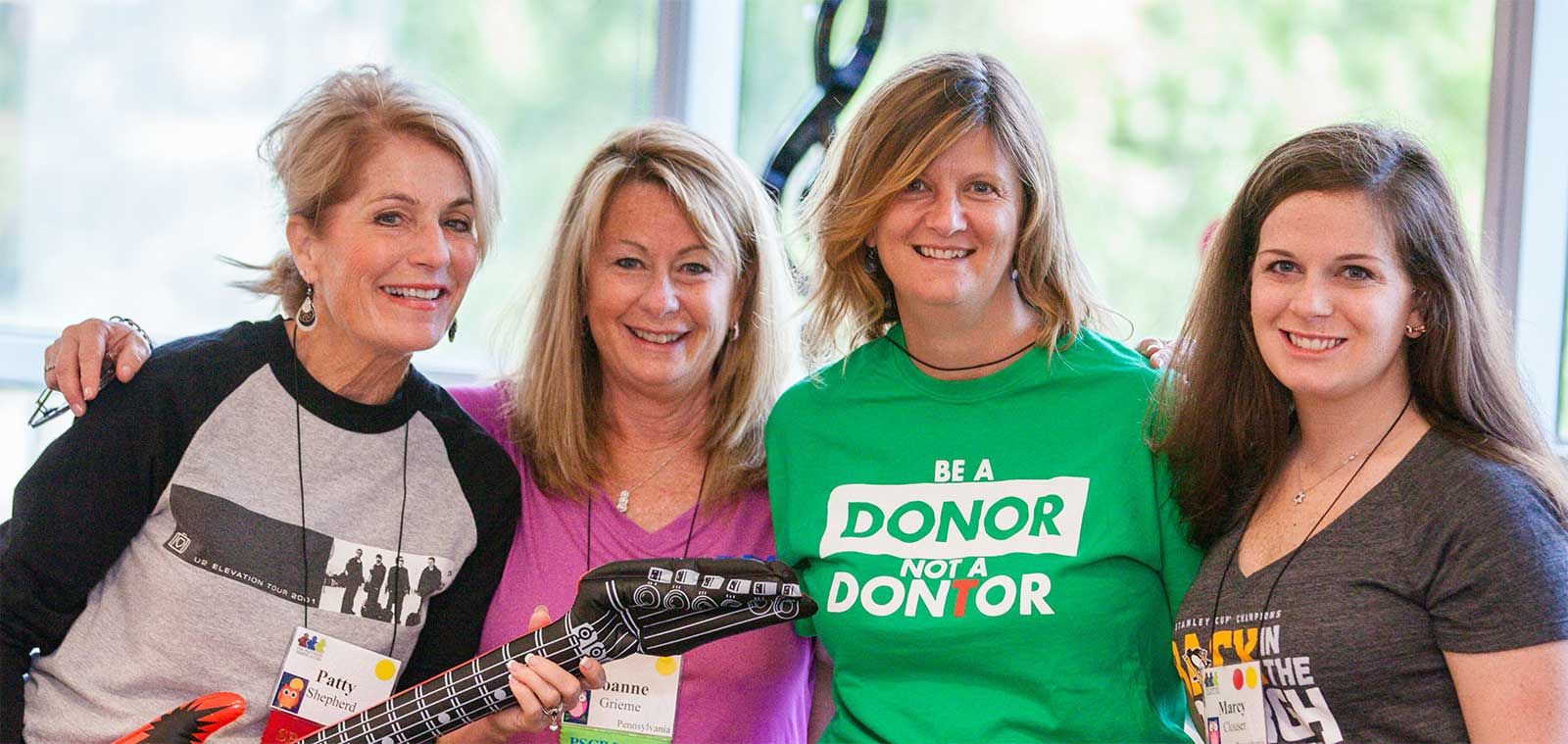 Four women stand in a line smiling at the camera. One woman is wearing a tshirt that say Be a Donor, Not a Don'ter