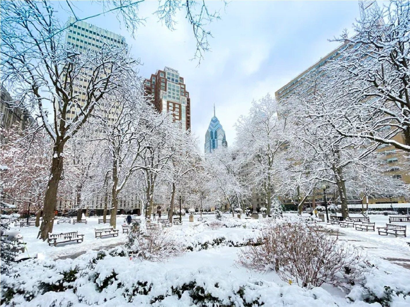 A park with trees and benches covered in snow with the Philadelphia skyline in the background.