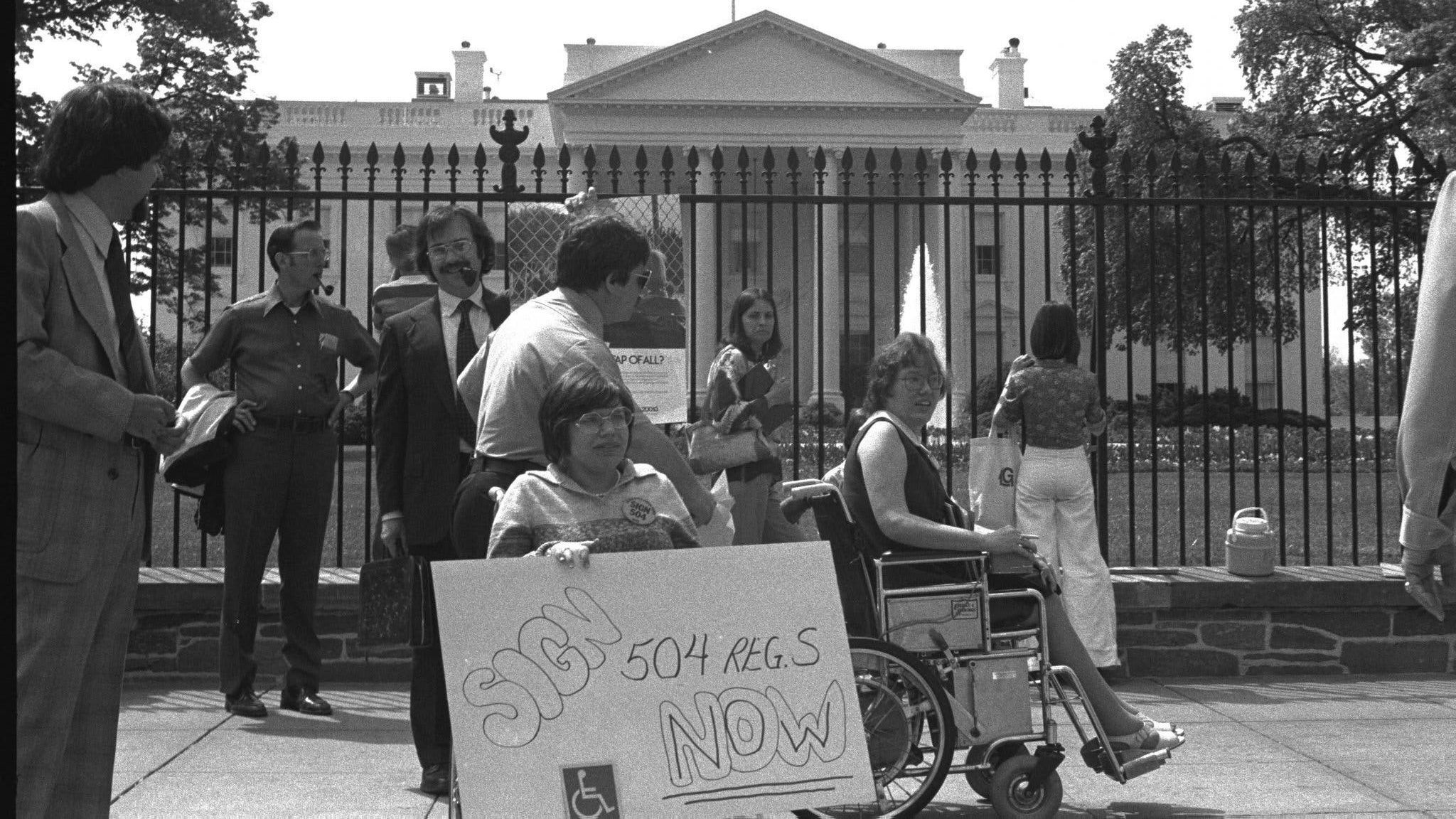 vintage black and white photo shows a group of disability rights activists, some in wheelchairs, with a protest sign in front of the White House