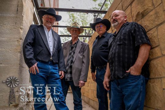 Performers for the Smoky Hill Museum Street Fair. Four men from the band Steel Skarecrow standing between two buildings.