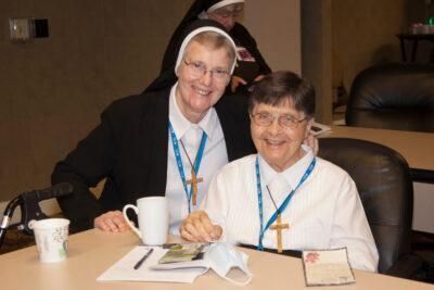 Two catholic sisters are seated at a table and are smiling at the camera. 
