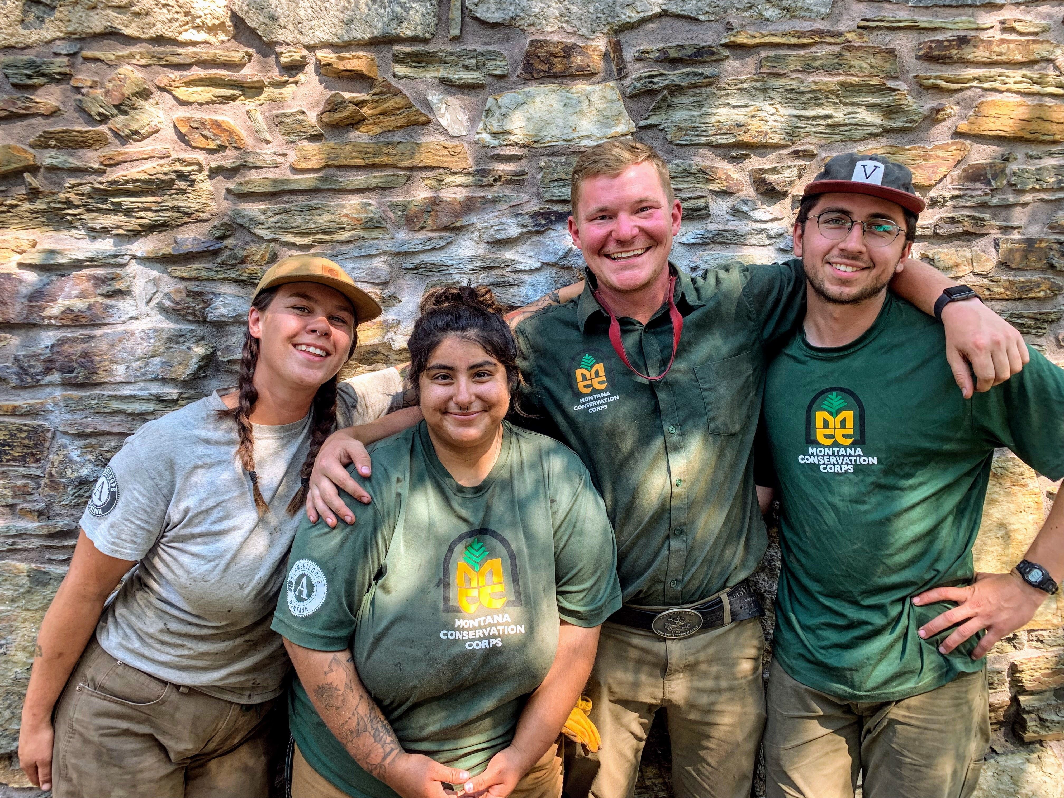 [Image Description: Four MCC members standing next to one another, smiling. They are all speckled with dirt from a project they were on. They are outside.]