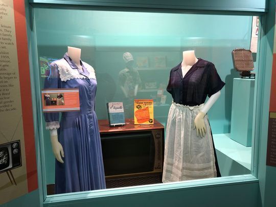 Exhibit case at the Smoky Hill Museum showing two 1950 dresses, a 1950 console TV, a Emily Post's Book of etiquette and an advertisement for a 1950s electric typritter 