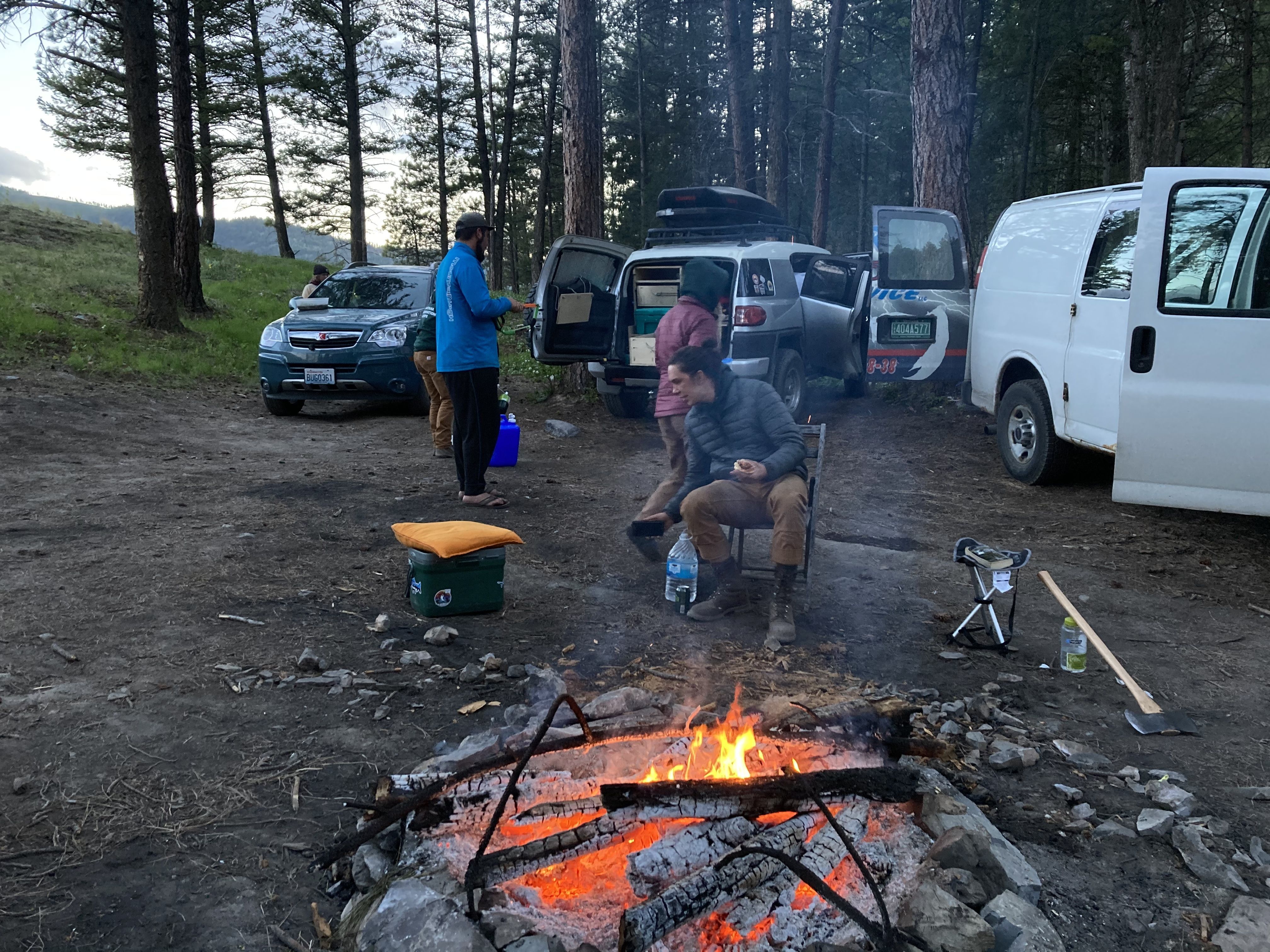 Crew members sit near a fire and walk around their camp, surrounded by vehicles they are living in.