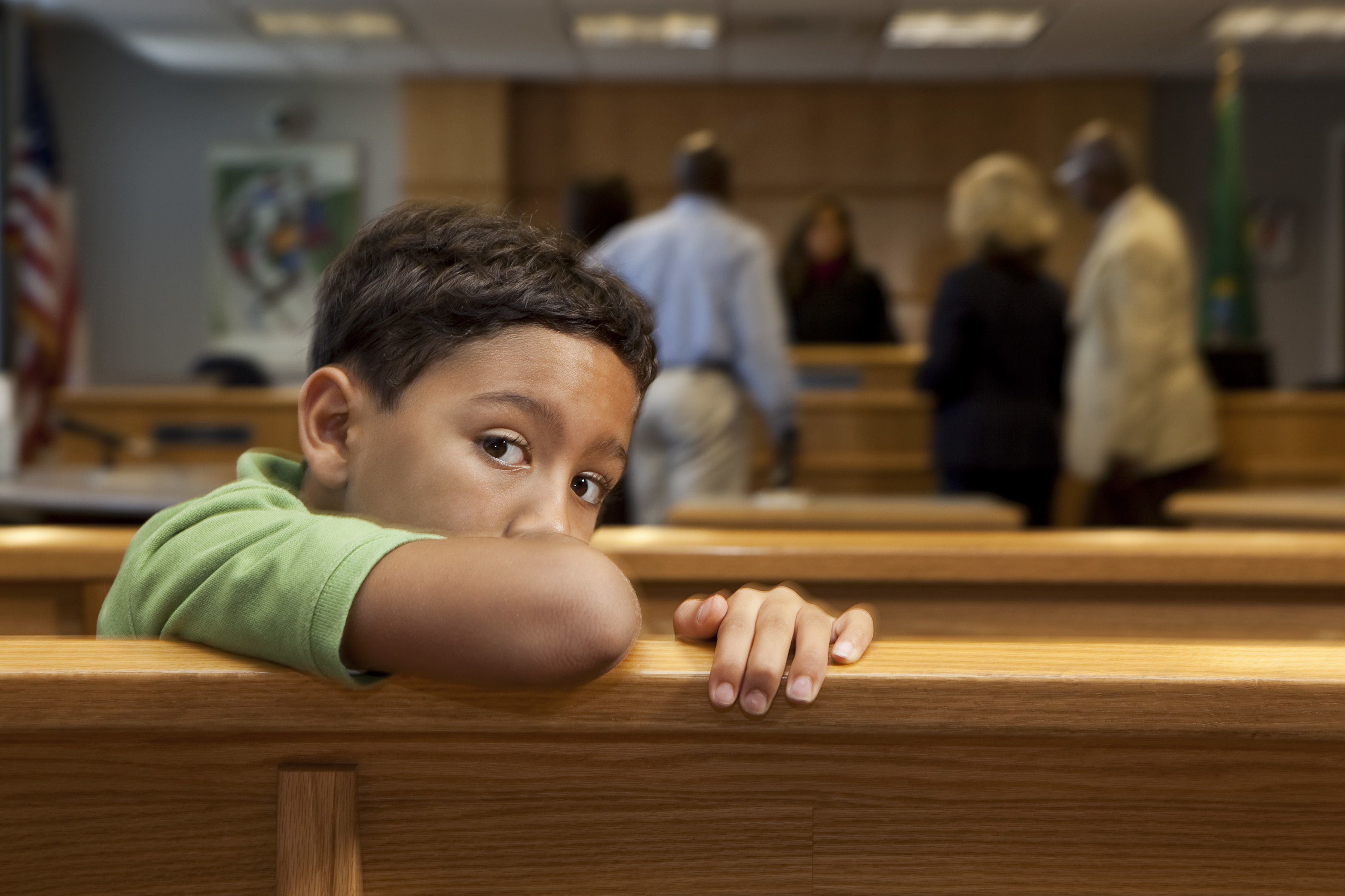 A boy sits on a bench in courtroom alone, his face looking over the back of bench at the camera.