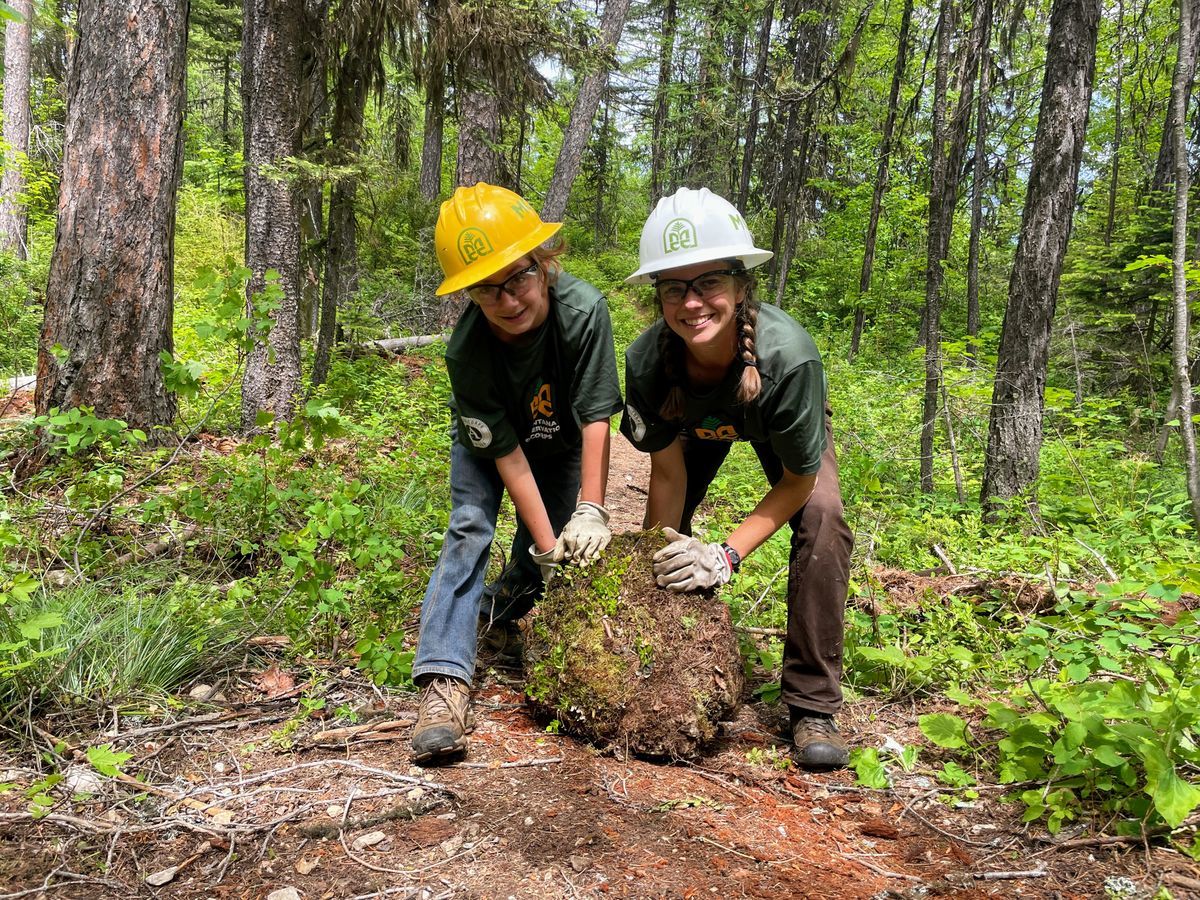 [Image Description: Two Youth Crew MCC members are shown holding what appears to be a large mound of tree trunk, standing in the middle of a forest.]