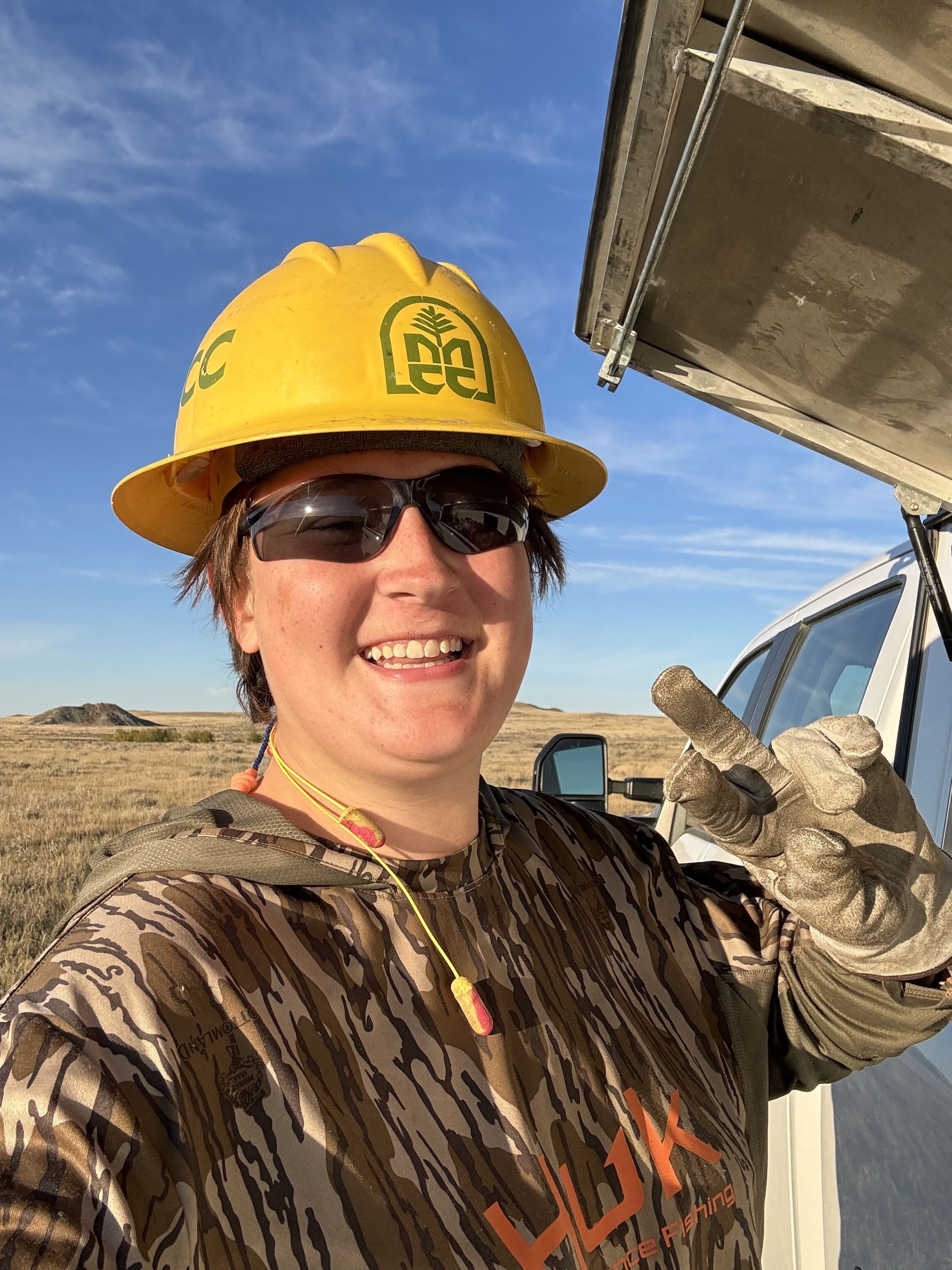 A close up of a crew member wearing a helmet, holding up a peace sign at the camera and smiling