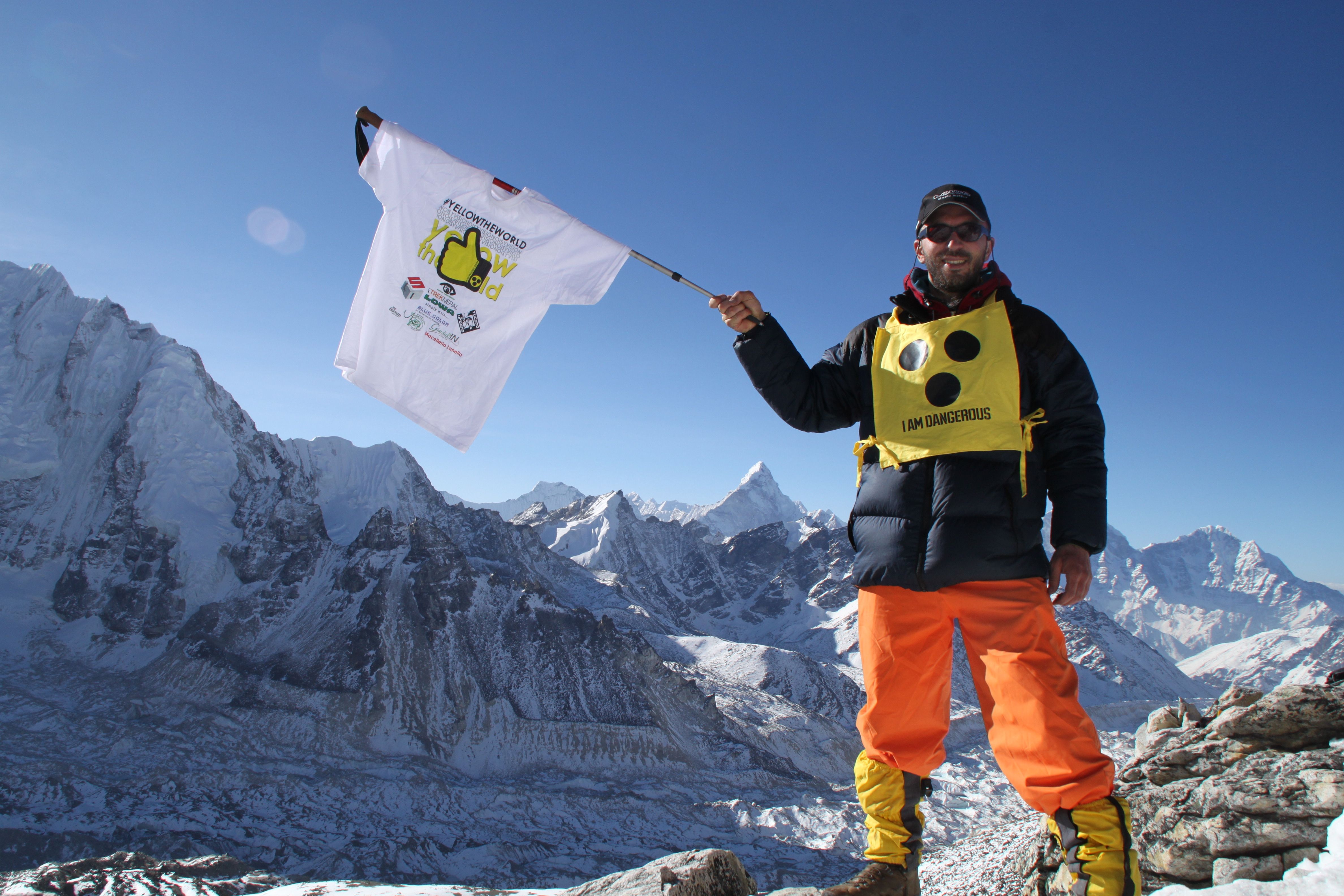 A photo of Dario Sorgato standing on top of a snow covered mountain holding a Yellow the World tee-shirt on a pole