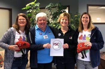 Four women, PSC Partners employees, stand smiling in front of a plant. They are holding stuffed livers and wearing PSC Partners tshirts.
