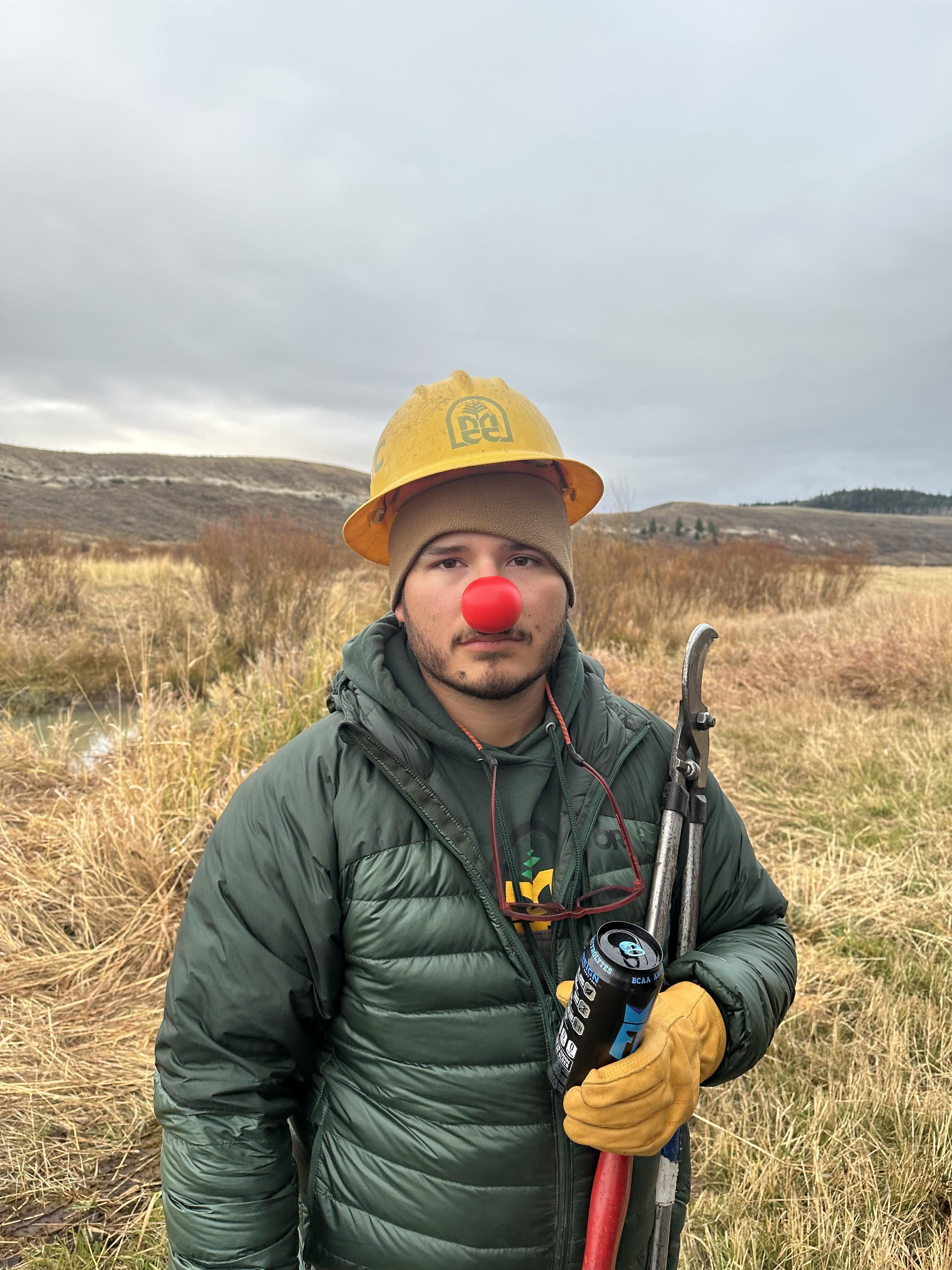 A crew member wearing a red nose and hard hat, holds a pair of loppers and stares into the camera.