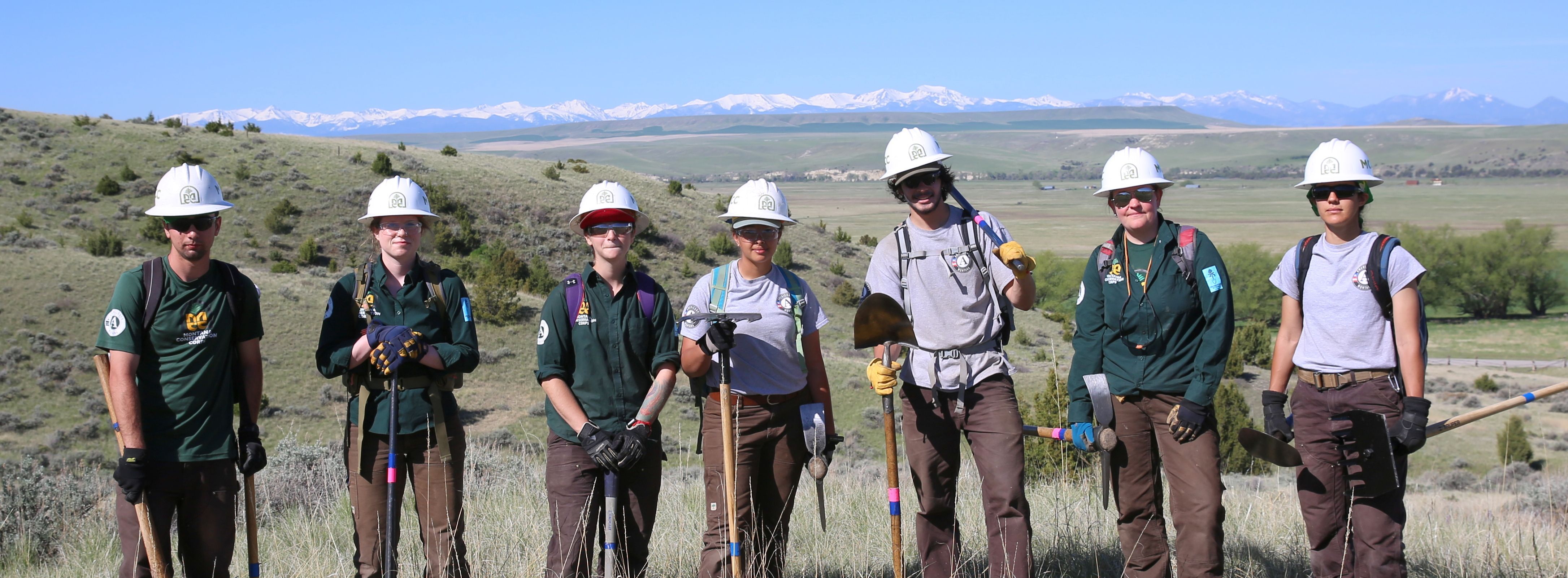 [Image Description: Seven MCC members holding their trail tools, donning their uniforms and hard hats. In the background is a mountain range, completely blanketed in snow.]