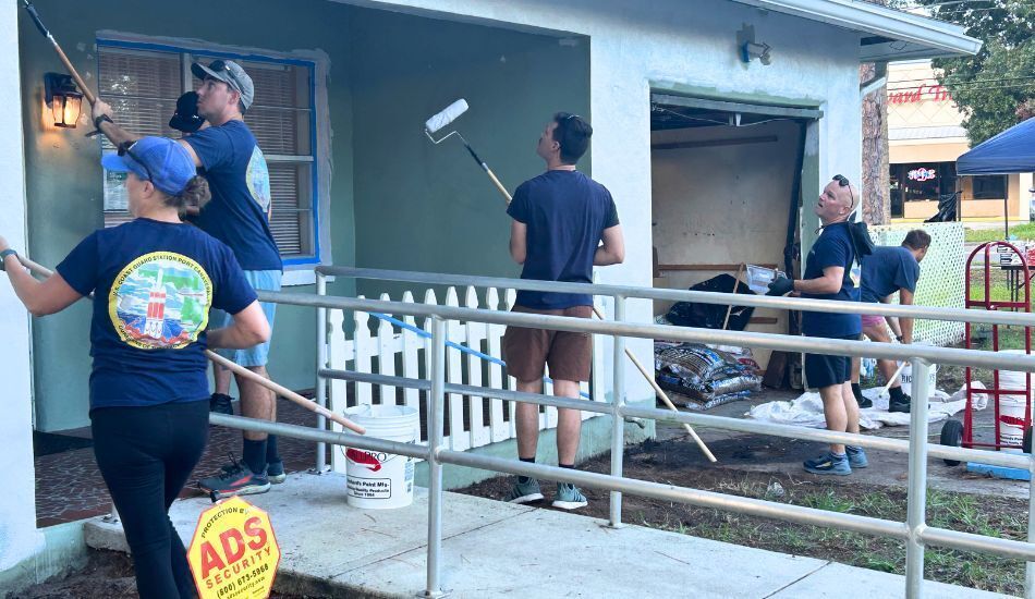 Photo of five volunteers painting the exterior of a home. They wear navy blue t-shirts and use rollers on long poles. The home was sage green and is being painted white.