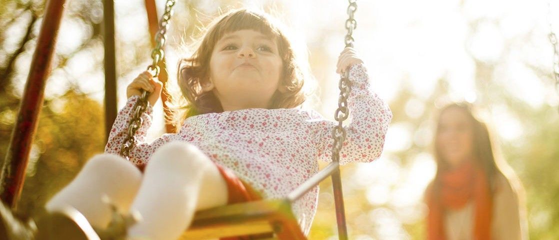 Photo of a young girl on a swing with her mother behind her.