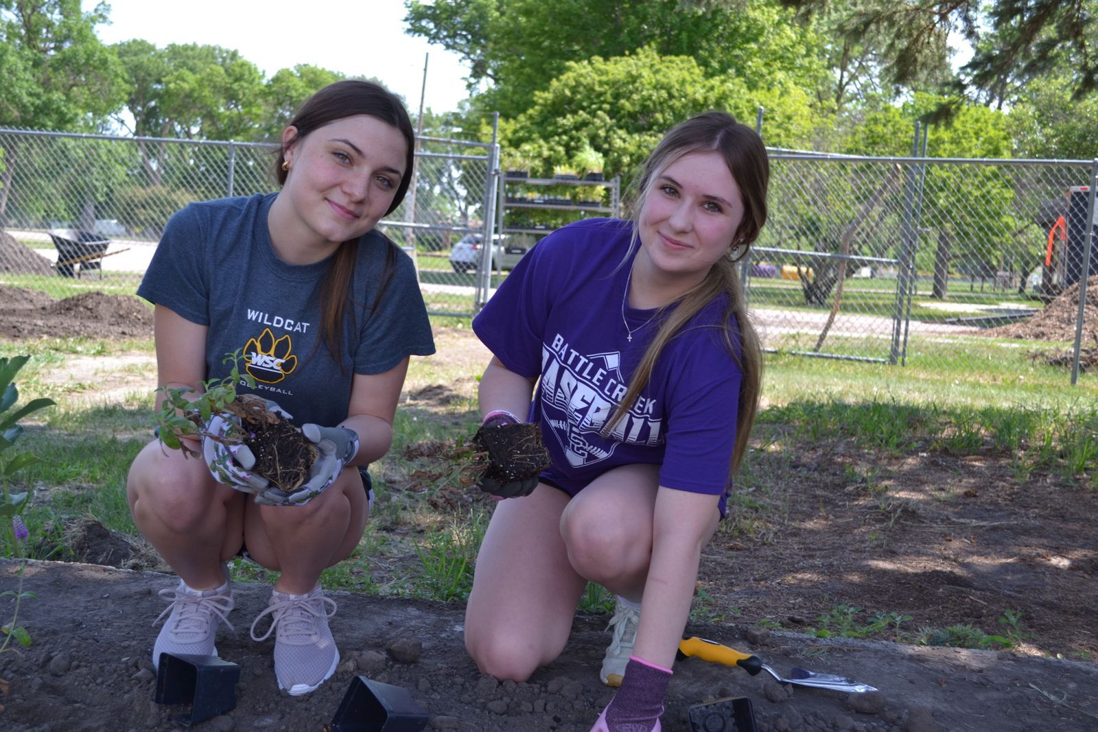 Two high school girls wearing purple tee shirts knee down to plant starter plants in a garden bed,