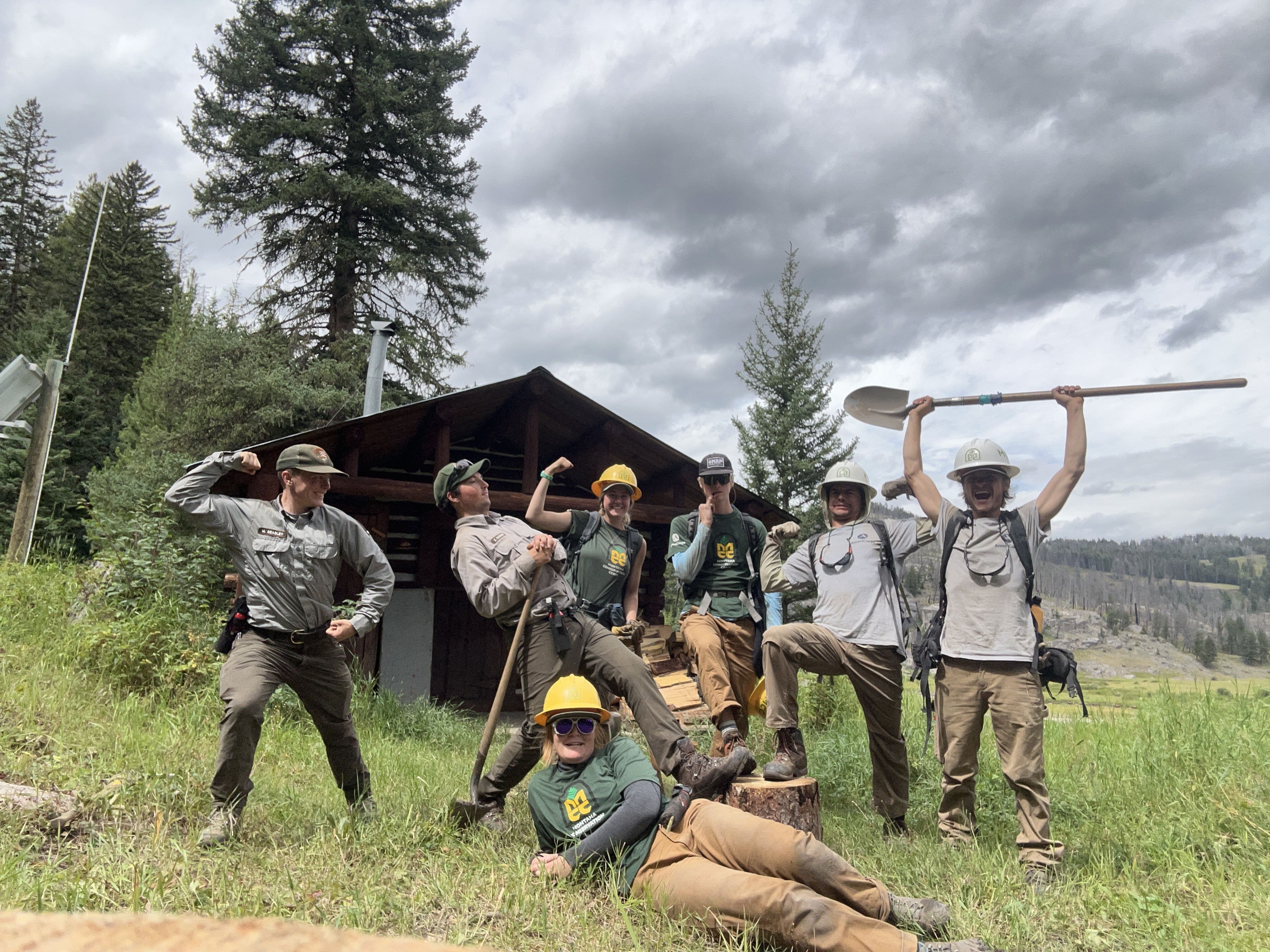 A crew and two National Park Service employees stand with tools in front of a cabin, making funny faces.