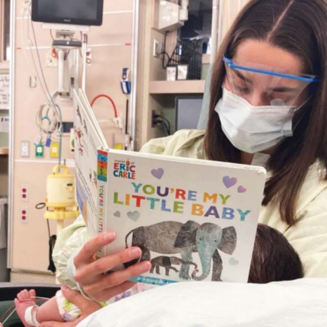 woman with mask, goggles, and hospital gown reads to baby in NICU