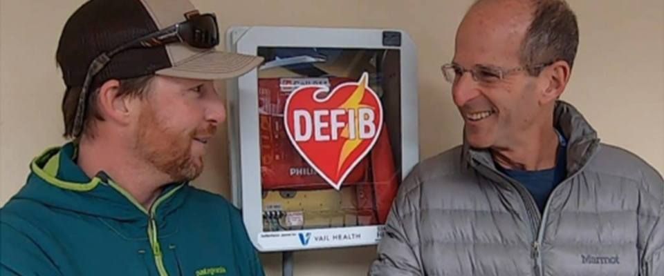 A photo of Doug Bartholomew and friend by a Defib machine.