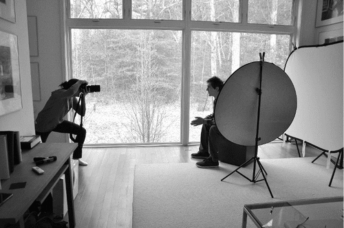 Black and white photo of a woman photographing a young man in a room with floor-to-ceiling windows and lots of light.