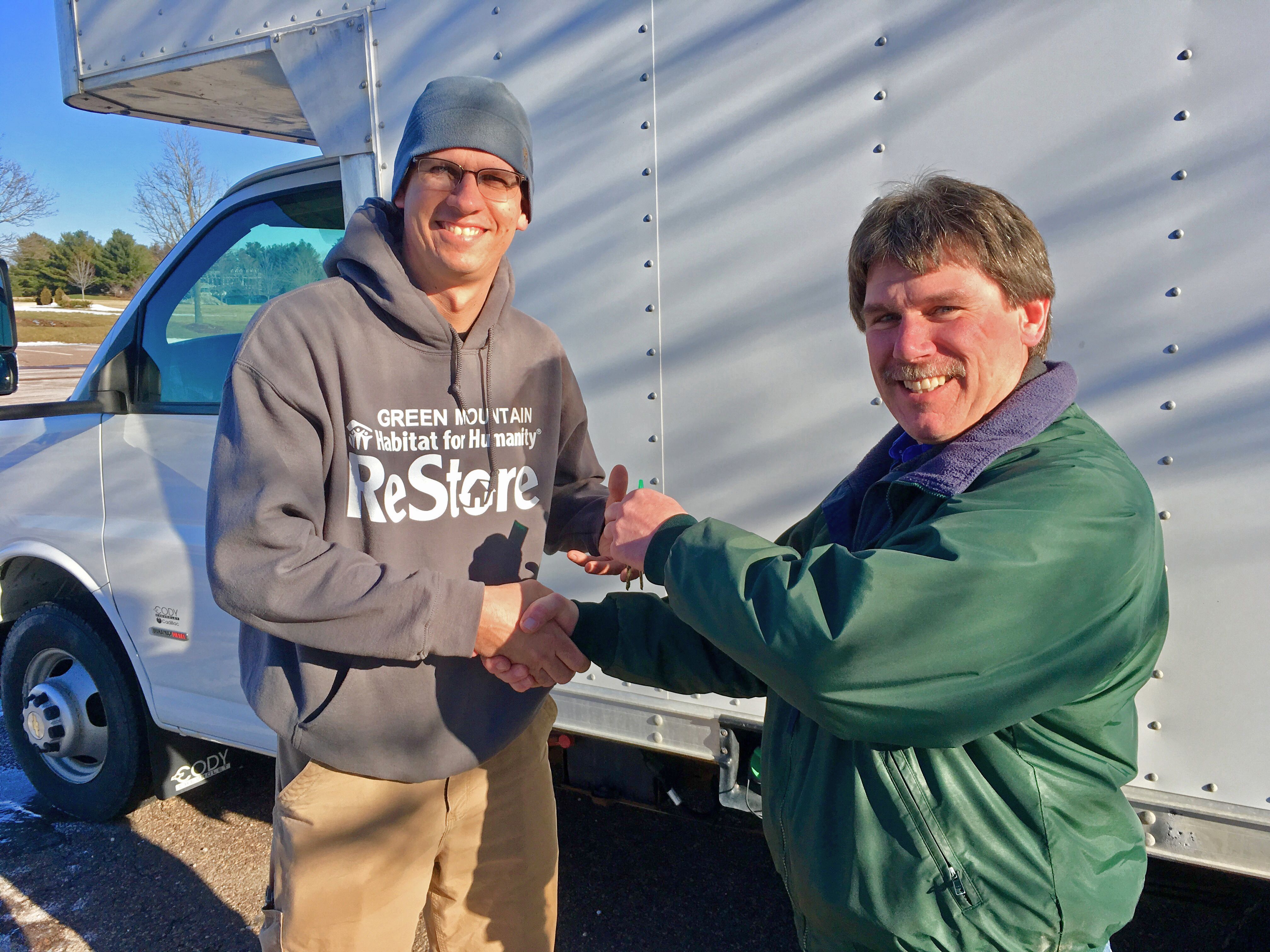 Two men shake hands in front of a truck.