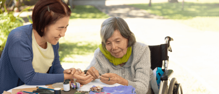 Woman with short dark hair smiles as she hands needlework to a woman with gray hair smiling and sitting in a wheelchair.