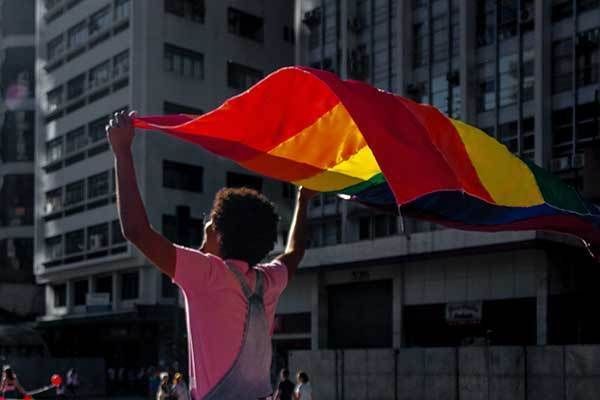 A person walking down the street while holding a rainbow flag with both hands overhead.